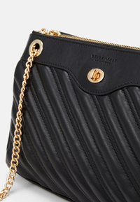 Mustassa tikatussa nahkalaukussa on kultaketjuhihna, kultaiset varusteet ja logo. Laukku on ohut ja suorakaiteen muotoinen.