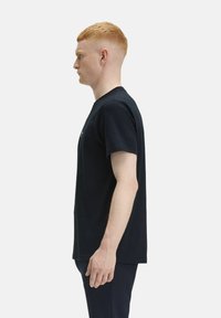 T-shirt nera in cotone a maniche corte con collo rotondo, esposta su un modello in profilo. Presenta un design semplice e privo di decorazioni.