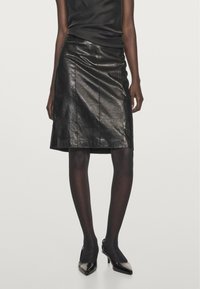 Vrouw die een zwarte, glanzende, gestructureerde rok tot op de knieën draagt, zwarte panty's en zwarte, puntige hoge hakken, tegen een egale achtergrond.