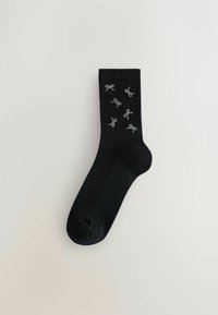Schwarze, gerippte Socke mit dekorativen silbernen Schleifen. Glatte Struktur, standardmäßige Sockengestaltung und verstärkter Zehenbereich.