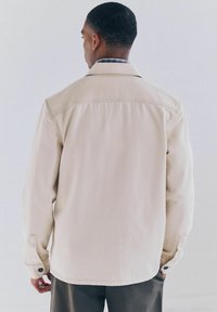 Beige katoenen jack met een ontspannen pasvorm, knoopmanchetten en een zacht gestructureerde afwerking. De achterkant toont minimale stiksels en een strakke uitstraling.