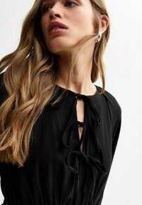 Black dress featuring a round neckline with ties, flowing fabric, and a fitted waist. Model has long, straight hair and wears a silver earring.
