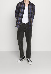 Chemise en flanelle à carreaux bleu marine et jaune, débardeur blanc, jean noir et baskets noires avec des touches blanches. Tenue simple et décontractée.