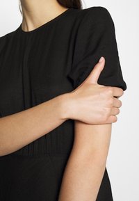Vestido negro con una tela texturizada, que presenta mangas cortas abullonadas y un corsé ajustado. Primer plano de una mano descansando sobre el brazo.