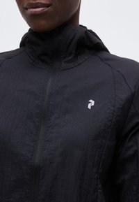 Κοντινό πλάνο ενός ατόμου που φοράει μαύρο μπουφάν με κουκούλα, μπροστινό φερμουάρ και μικρό λευκό γεωμετρικό λογότυπο στο στήθος.