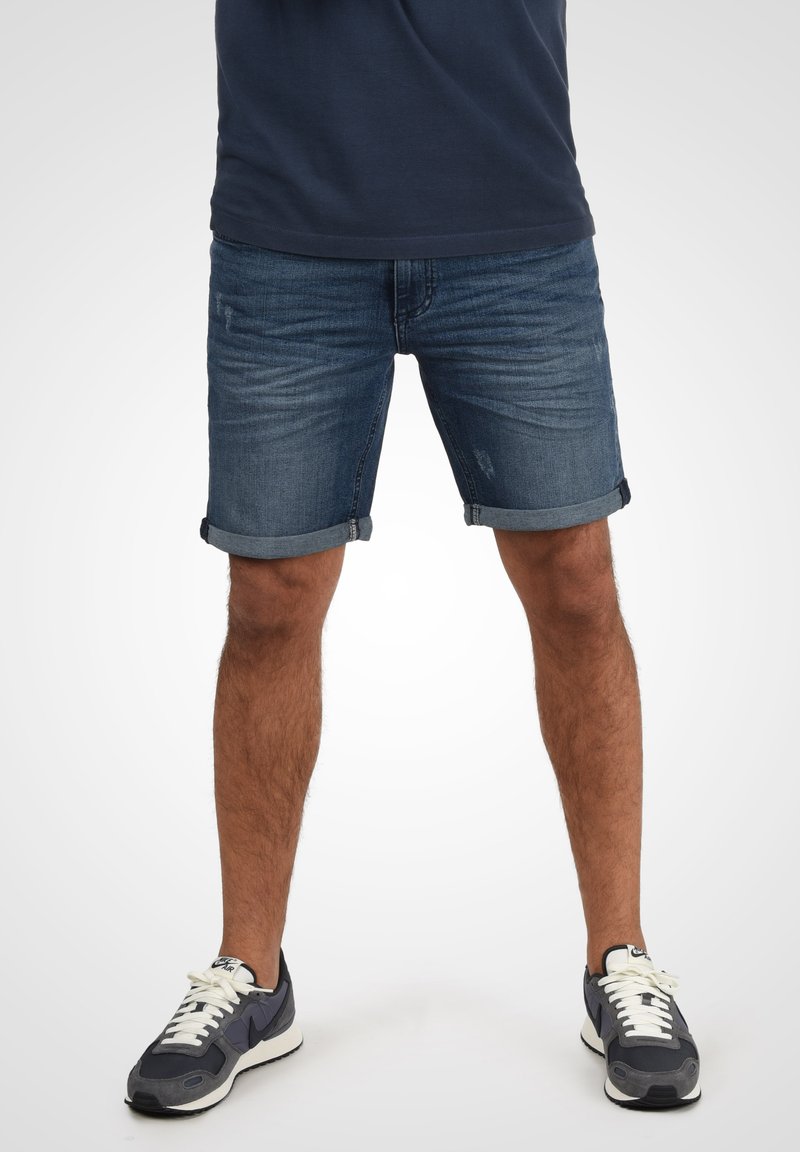 Jeans shorts i mørkeblå med optrukne kanter, der har en afbleget tekstur og subtile slidmærker, parret med grå sneakers og hvide snørebånd.