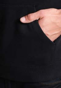 Černá mikina s žebrovaným lemem, boční kapsou a měkkou texturou. Částečně viditelná ruka spočívá v kapse.