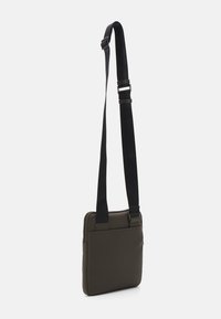 Sac à dos en vert olive foncé, fabriqué en matériau lisse, doté d'une longue sangle noire réglable et d'une fermeture zippée.