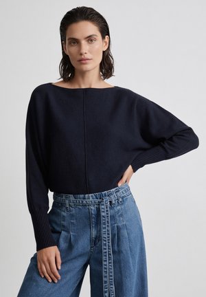 Frau mit kurzen dunklen Haaren, die einen lockeren schwarzen Pullover und hoch taillierte blaue Jeans mit Gürtel trägt, steht mit einer Hand in der Hüfte.