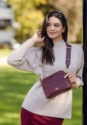 Bordeaux kožená kabelka přes rameno s prošívaným vzorem, zlatými kovovými doplňky a nastavitelným popruhem, nošená modelem na zeleném pozadí.