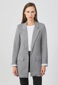 Blazer grigio realizzato in tessuto morbido, con colletto a punta, due tasche frontali e accenti a righe sui polsini. Indossato sopra un top bianco.