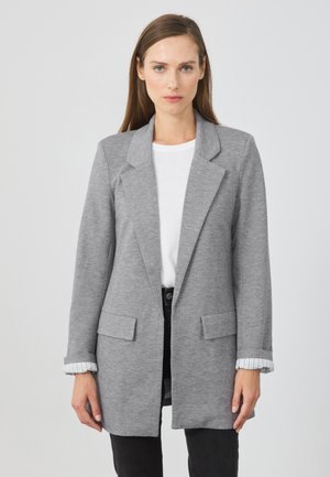 Blazer gris en tissu doux, avec un col cranté, deux poches à l'avant et des accents rayés aux poignets. Porté par-dessus un top blanc.