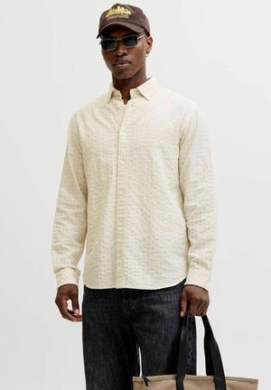 Homme portant une chemise beige texturée boutonnée, un jean foncé, une casquette marron, des lunettes de soleil et tenant un sac fourre-tout beige avec des poignées noires.