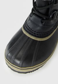 Botín de invierno negro impermeable con punta de goma estriada, ribete beige y cordones negros sobre fondo blanco.