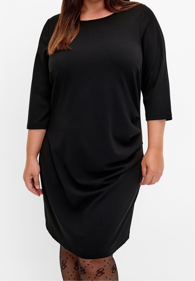 Femme portant une robe noire longueur genou avec des manches trois-quarts et des collants noirs transparents avec un motif de nœud et de points.