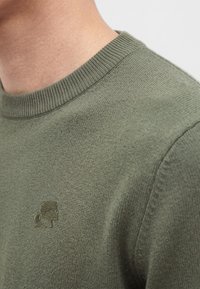 Gros plan d'une personne portant un pull en maille vert olive avec un petit logo brodé sur la poitrine près du col.