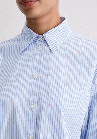 Light blue and white striped button-up shirt with a pointed collar, featuring a chest pocket and white buttons along the placket.