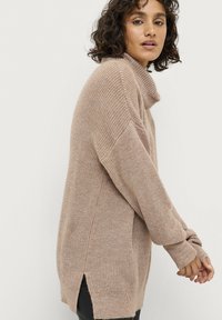 Overdimensioneret beige strikket sweater med høj hals, ribbet tekstur, sidelæg og lange ærmer. Blød, afslappet pasform, der er velegnet til lag-på-lag.