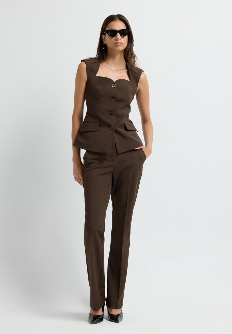 Gilet marron cintré avec un décolleté carré et une fermeture par boutons, associé à un pantalon ajusté assorti et des escarpins noirs à bout pointu.