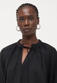 Black blouse with a gathered neckline, tie detail at the collar, and a v-shaped opening. Made of lightweight fabric with a smooth texture.