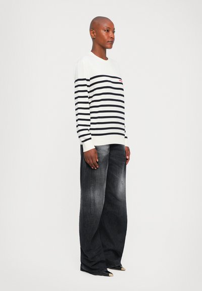 Person standing wearing white and black striped sweater and wide-leg faded black jeans with pointed-toe black shoes.