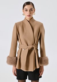 Cappotto avvolgente tan in tessuto texturizzato, con vita accentuata da cintura e morbidi polsini in pelliccia. Presenta un colletto sagomato e una chiusura a doppi bottoni per un aspetto strutturato.