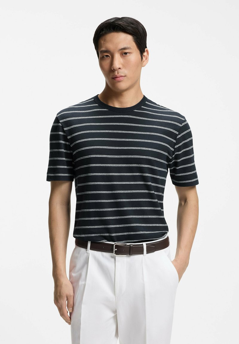 Homme portant une chemise à manches courtes rayée noir et blanc, un pantalon blanc et une ceinture marron, debout avec une main dans la poche sur fond blanc.