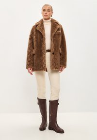 Brauner Faux-Fur-Mantel mit strukturiertem Muster, lässigem Schnitt und zwei vorderen Taschen. Getragen über einem cremfarbenen Rollkragenpullover und kombiniert mit braunen kniehohen Stiefeln.