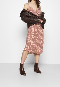 Veste matelassée en simili cuir marron sur une robe midi rose à pois blancs. Bottines marron à finition texturée. Tissu lisse.