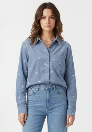 Femme aux cheveux bruns ondulés portant une chemise bleue claire brodée à boutons rentrée dans un jean bleu clair taille haute, debout sur un fond blanc.