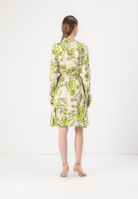 Rochie florală cu fundal bej și modele verzi vibrante, mâneci lungi, talie strânsă și fustă evazată. Sandale cu toc înalt, transparente.