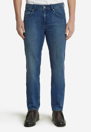 Blaue Denim-Jeans mit geradem Schnitt, Fünf-Taschen-Design, Metallknopf und Nieten sowie dezentem Verblassen. Getragen mit schwarzen Schuhen.