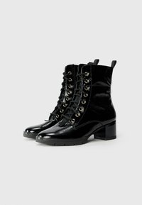 Botas de tobillo de charol negro con tacón cuadrado, que cuentan con ojales en tono dorado, cordones negros y dos asas en la parte superior para facilitar su uso.