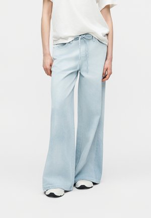 Person iført lyseblå vidde cowboybukser med bundet talje og hvide sneakers, stående mod en ensartet hvid baggrund.