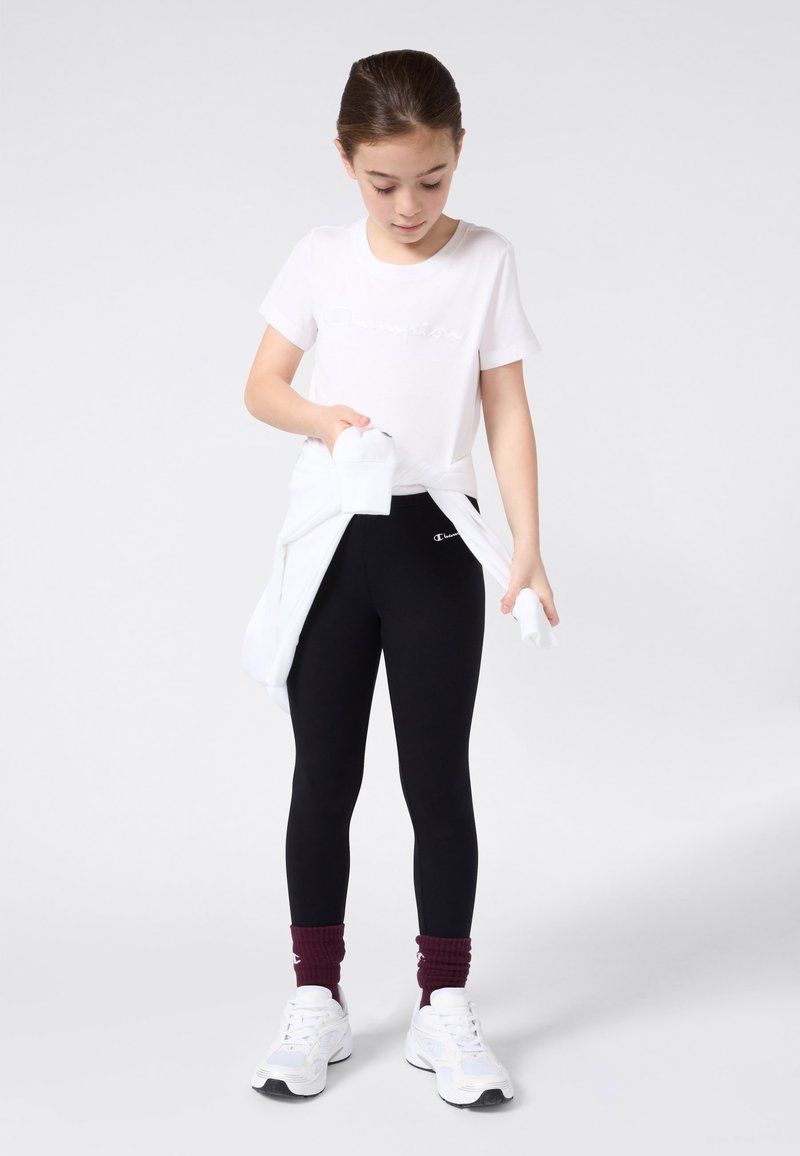 Witte katoenen t-shirt met reliëf "Champion" logo, zwarte leggings, witte sport schoenen en bordeauxrode crew sokken. Witte trui om de taille geknoopt.