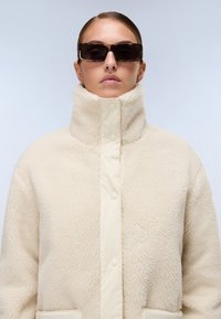 Casaco de polar creme com colarinho alto, apresentando botões de pressão e uma textura suave. Óculos de sol retangulares escuros completam o visual.