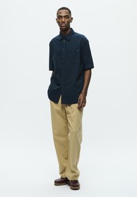 Cămașă scurtă cu mânecă, de culoare bleumarin închis, cu un buzunar pe piept, asociată cu pantaloni lejeri de culoare bej deschis și pantofi maro slip-on.