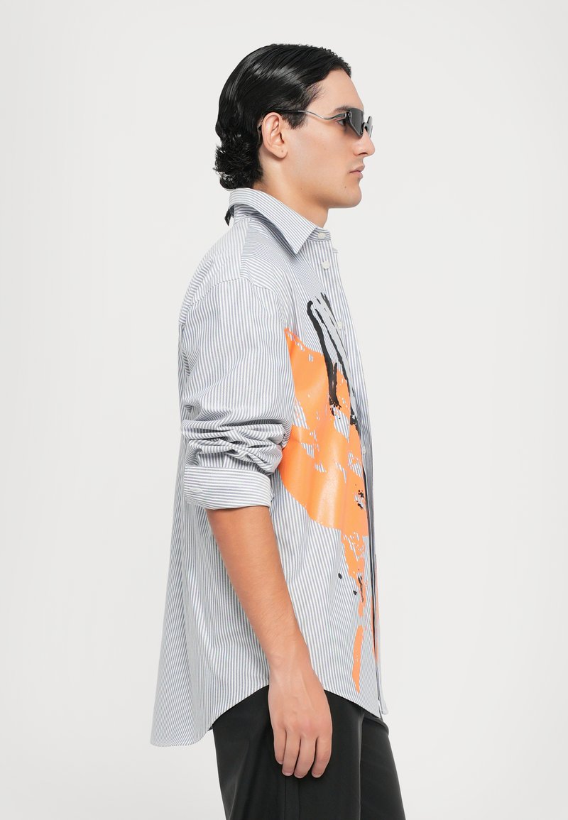 Gestreiftes Hemd mit knöpfen, das ein orangefarbenes abstraktes Design auf der Vorderseite aufweist, mit aufgerollten Ärmeln und einer lockeren Passform. Getragen mit schmalen schwarzen Hosen.