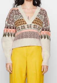 Kvinna i en flerfärgad mönstrad V-ringad tröja och ljusgula högmidjade byxor, stående med händerna avslappnat vid sidorna.