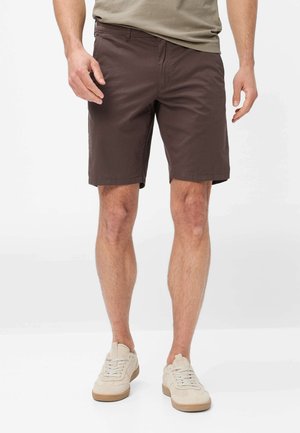 Mand iført mørkebrune knælange shorts og beige sneakers, stående mod en ensfarvet lys baggrund.