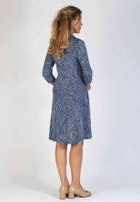 Niebieska sukienka w wzór paisley, z długimi rękawami, długości do kolan i z guzikiem na rękawach. Noszona z jasnymi, blokowymi obcasami.