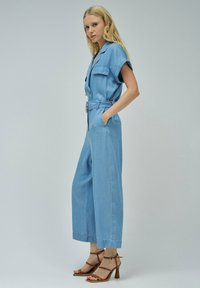 Mono de denim en azul claro, mangas cortas, con botones en el frente, cinturón en la cintura, dos bolsillos en el pecho, silueta de pierna ancha, combinado con tacones marrones de tiras.