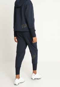 Persona vistiendo una sudadera oscura de color navy y joggers a juego con detalles en cursiva, combinados con zapatillas deportivas blancas, de pie sobre un suelo claro.