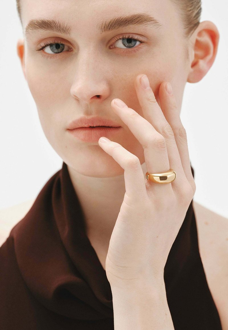 Anillo de oro con un diseño ovalado y suave, llevado en un dedo. El modelo tiene una expresión neutral y lleva una parte superior de tela suave y oscura.