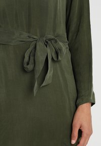 Gros plan d'une robe verte à manches longues avec une ceinture en tissu nouée à la taille et une main reposant à côté.