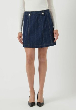 Kvinde iført en mørkeblå denim A-linjeet mini nederdel med lodrette syninger og sølvknapdetaljer langs taljen.