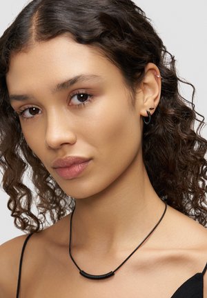 Schwarzes Halsband mit einem gebogenen Metallakzent und glatter Textur. Lockiges Haar und Creolen ergänzen den Look.