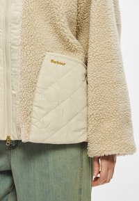 Beežist fliisjakk tikitud kreemjas taskuga, mille peal on kuldsete tähtedega kiri "Barbour". Jakil on tõmbluku sulgemine ja laiad, pehmed varrukad.