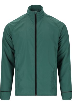 Person trägt eine grüne Jacke mit Reißverschluss, schwarze Shorts, schwarze Socken und graue Laufschuhe, steht mit verschränkten Händen auf der Taille.