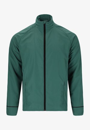 Personne portant une veste verte zippée, un short noir, des chaussettes noires et des chaussures de course grises, debout, les mains jointes à la taille.